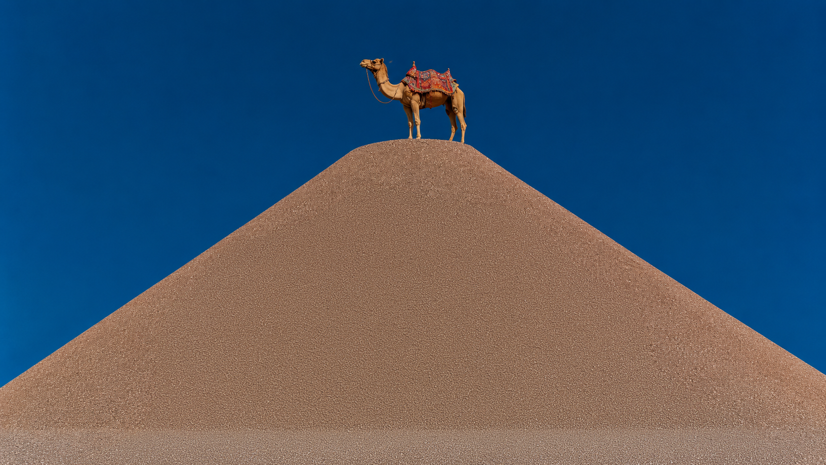 A camel standing on a large mound against a clear blue sky